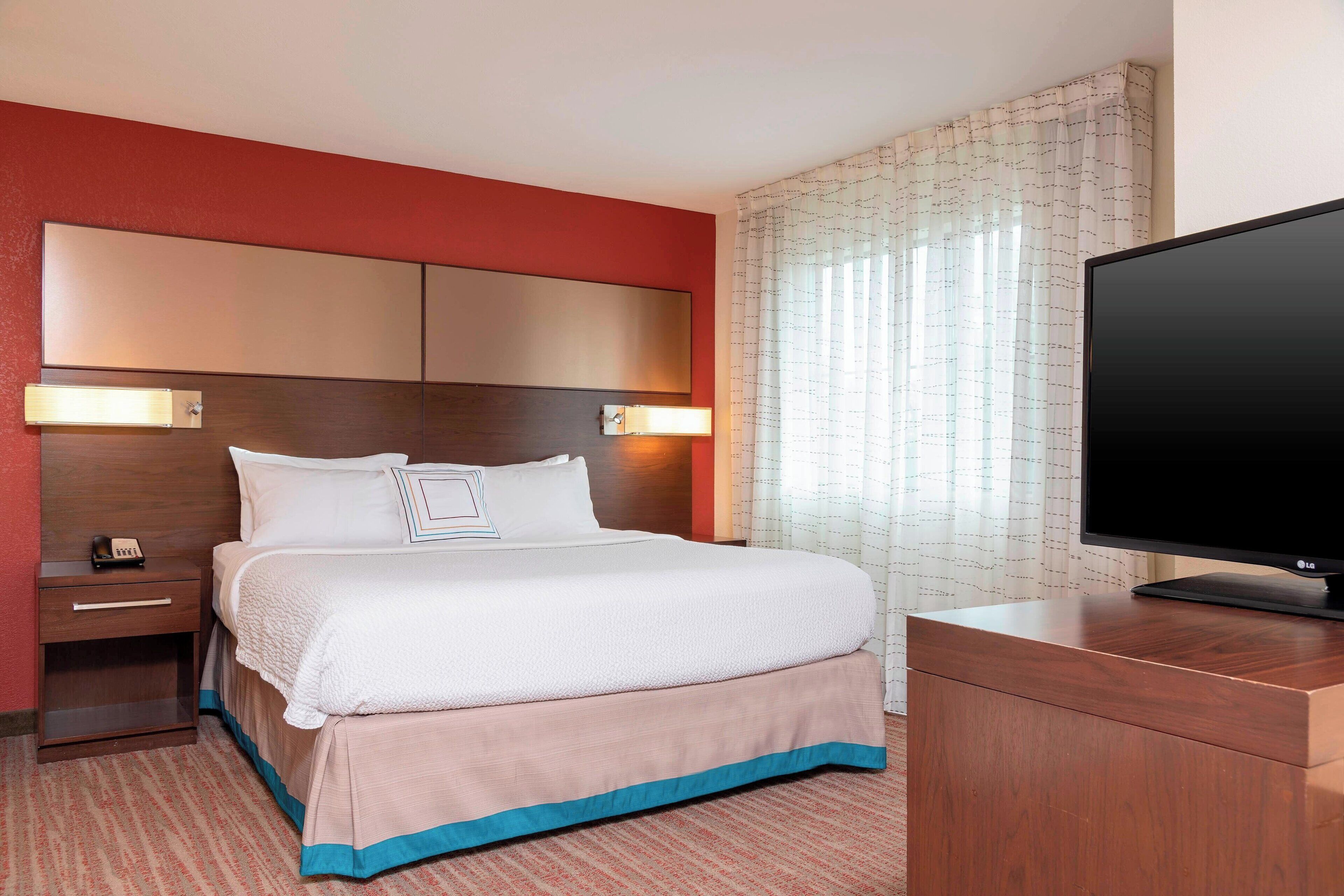 Een hotelkamer met een groot bed, een nachtkastje van hout, een flatscreen televisie en een raam met dunne gordijnen.
