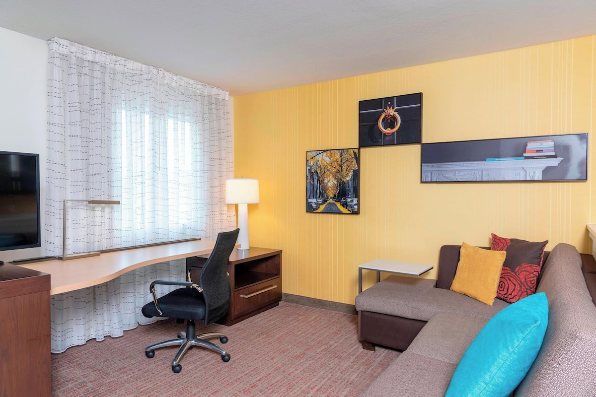 A hotel room with a desk, a TV, a sofa, and two wall-mounted artworks.