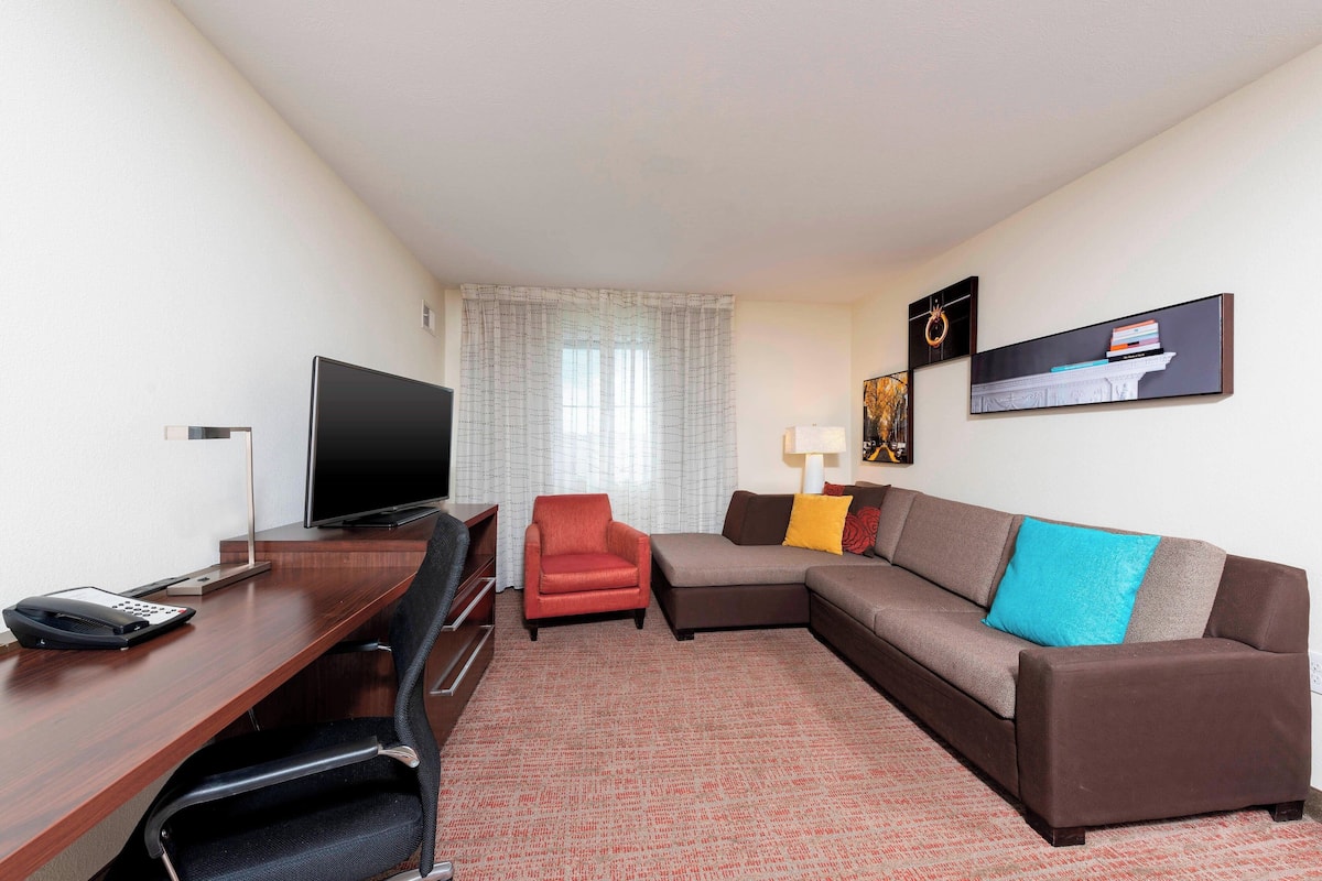 A hotel room with a desk, a television, a red armchair, a brown sofa with a blue pillow, and a wall-mounted artwork.
