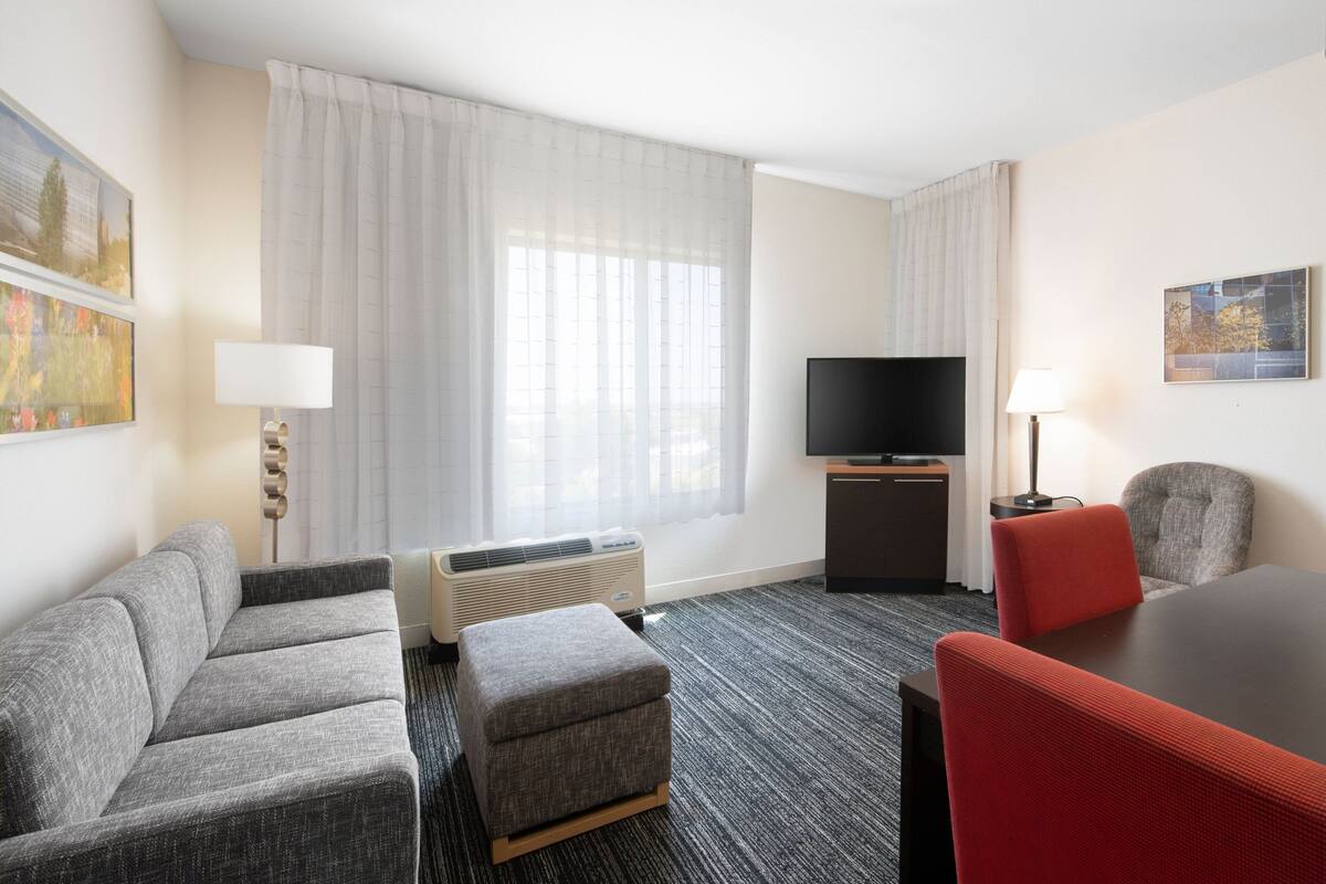 Ein Hotelzimmer mit grauem Sofa, einem roten Sessel, einem kleinen Hocker, einem Flachbildfernseher und einem Heizgerät.