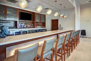 A bar area with a long counter, multiple bar stools, a television, and various bottles and glasses. - Springhill Suites Dallas Plano/Frisco (Plano)