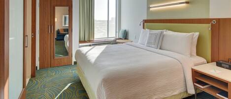 A hotel room with a large bed, a bedside table, a nightstand, a window with curtains, a mirror, and a wooden wardrobe.