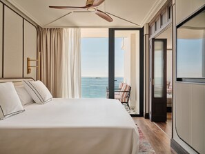 A hotel room with a large bed, a ceiling fan, a view of the sea, and a balcony with a chair.