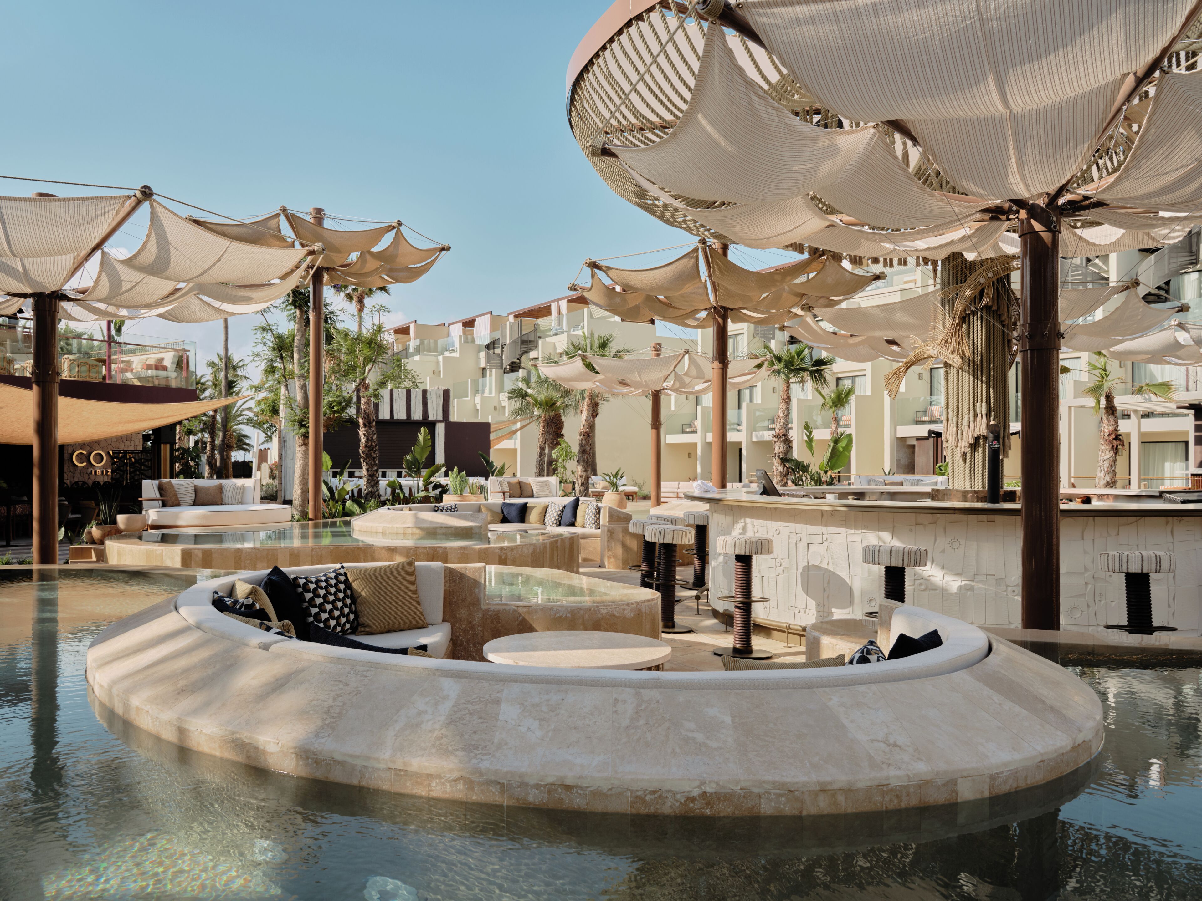 A pool area with lounge chairs and umbrellas, surrounded by palm trees and a building with balconies.