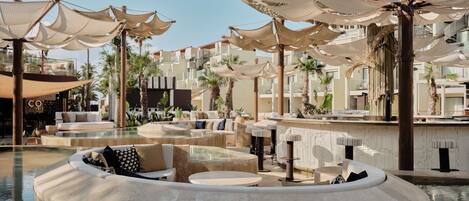 A pool area with lounge chairs and umbrellas, surrounded by palm trees and a building with balconies.