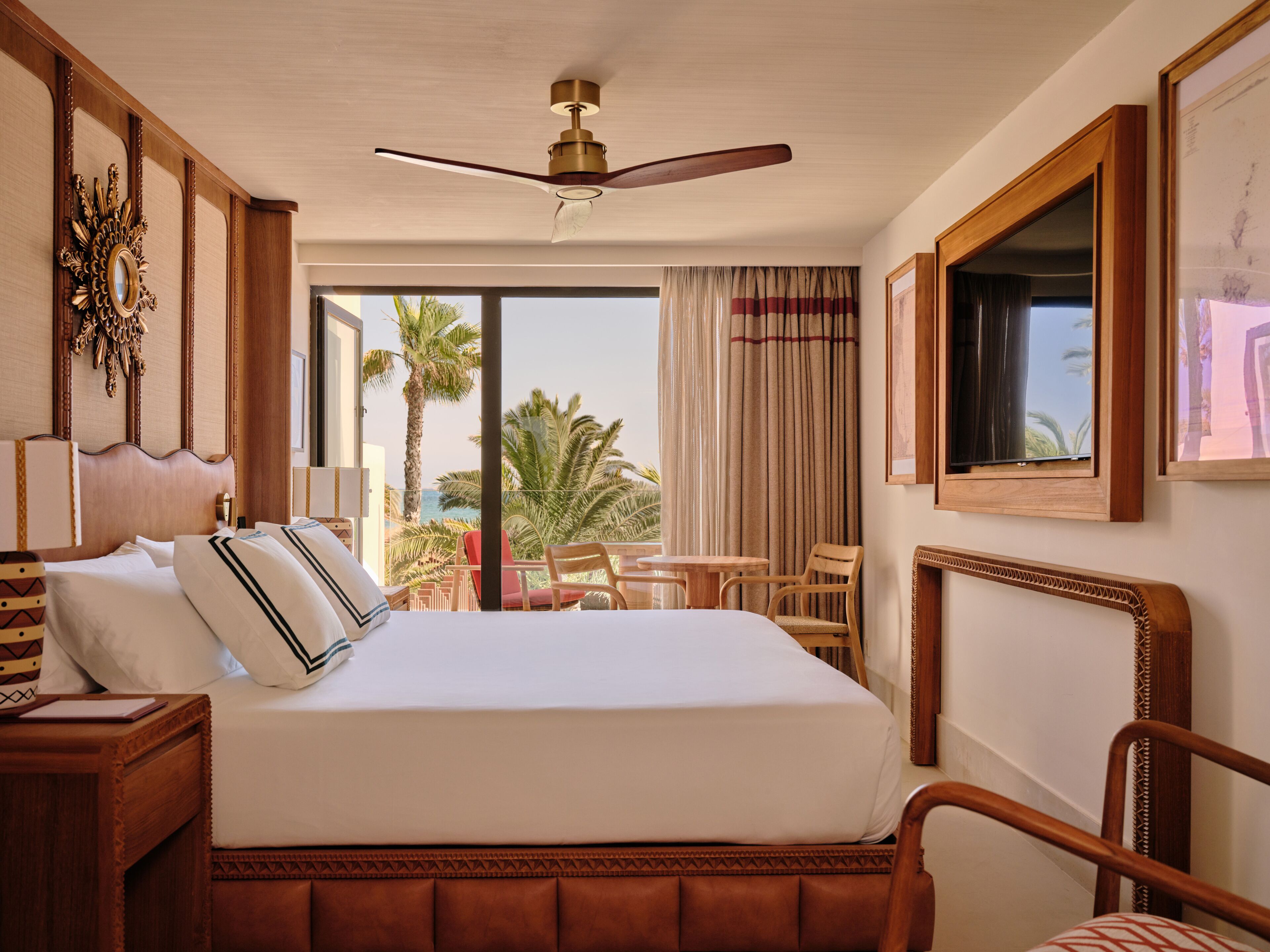 A hotel room with a large bed, a ceiling fan, a view of palm trees, and a balcony with chairs.