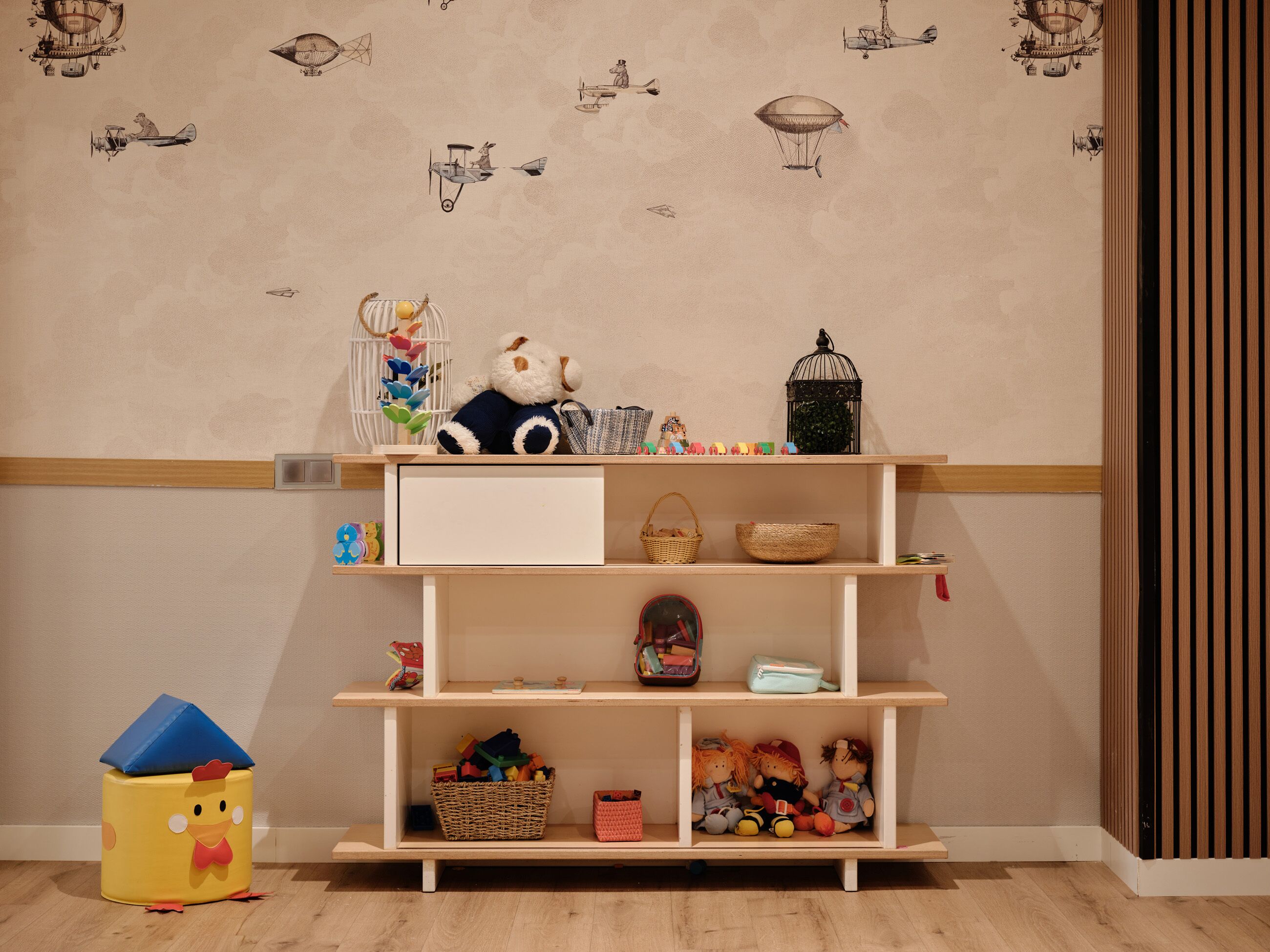 Ein Zimmer mit Holzboden, einem weißen Regal mit Körben und Spielzeug, und einer Wand mit Flugzeugmotiven.