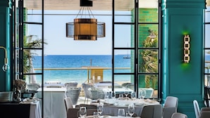 Un comedor con mesas dispuestas para una comida, ventanas amplias que ofrecen vista al océano y una lámpara colgante.