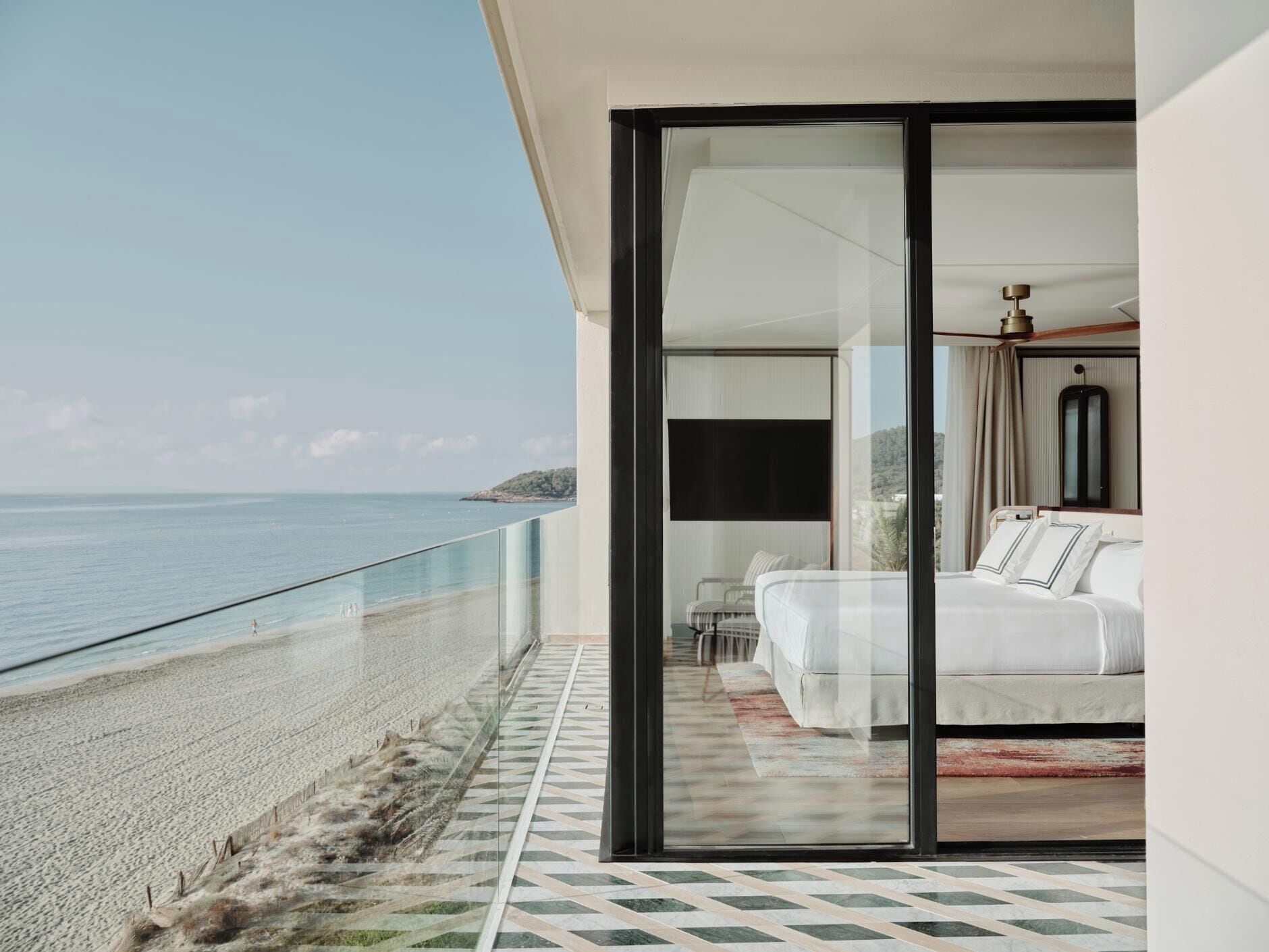 A modern balcony with a glass door leading to a bedroom with a bed and a chair. The balcony overlooks a beach and the sea.