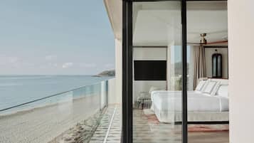 A modern balcony with a glass door leading to a bedroom with a bed and a chair. The balcony overlooks a beach and the sea.