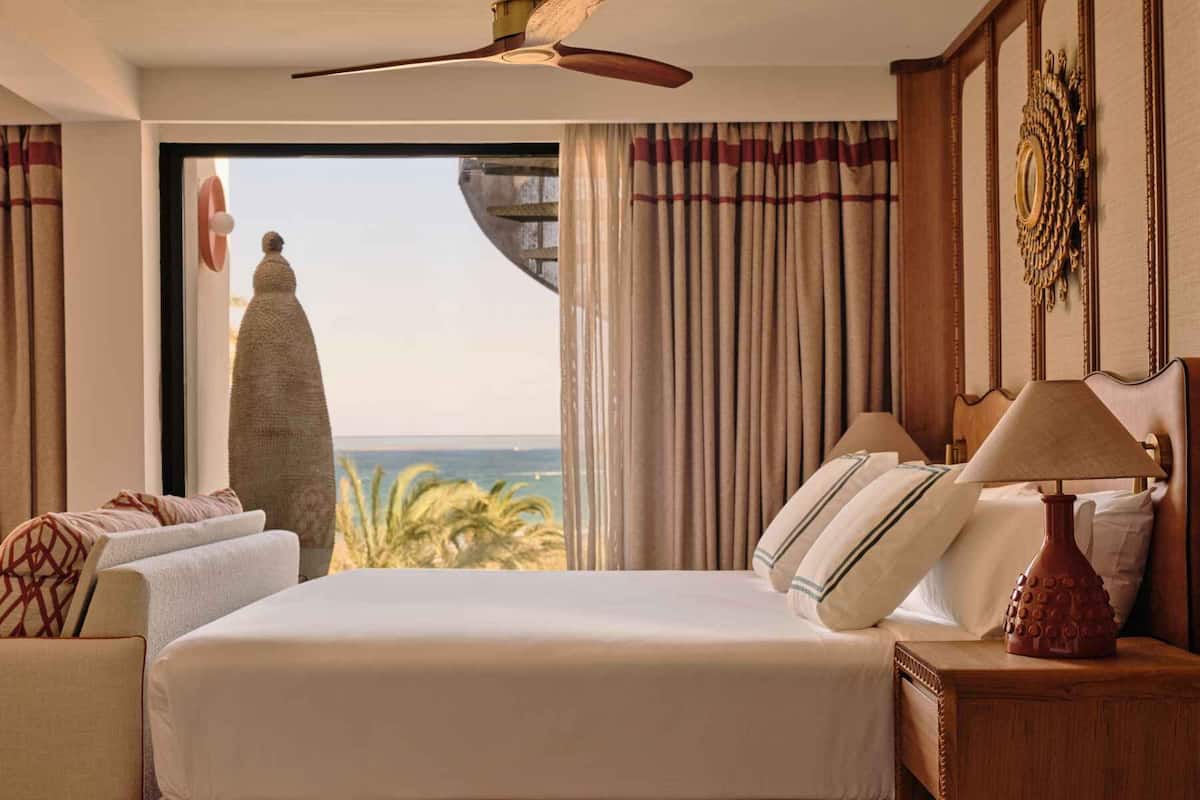 Ein Hotelzimmer mit Bett, Nachttisch, Lampe, Deckenventilator und Meerblick.