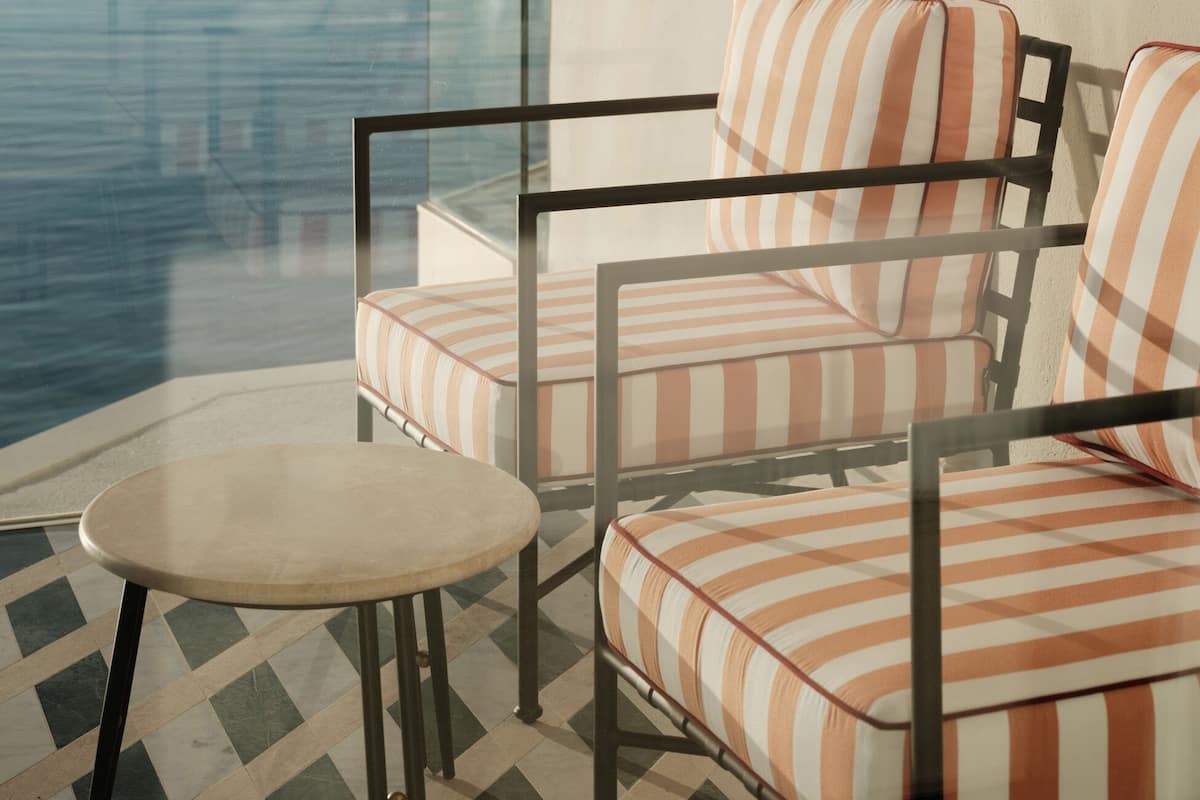 A balcony with a glass railing, two striped cushioned chairs, a small round table, and a view of the sea.