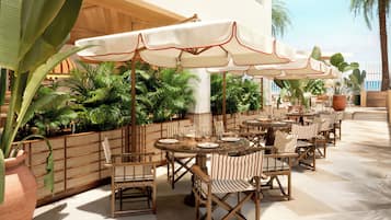 Un área de comedor al aire libre con varias mesas y sillas, rodeada de macetas con plantas y sombrillas.