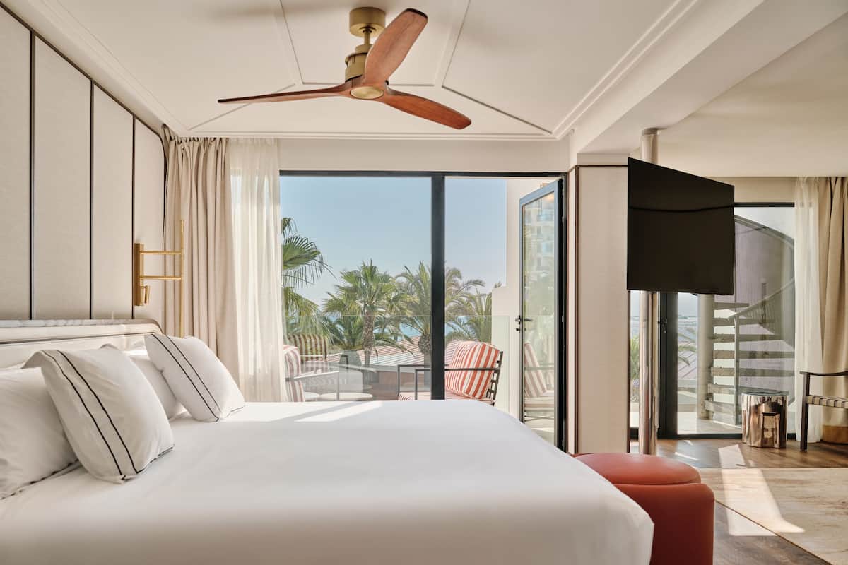 Ein modernes Hotelzimmer mit einem großen Bett, einem Deckenventilator, einem Balkon mit Blick auf Palmen und einer Treppe, die in einen Außenbereich führt.