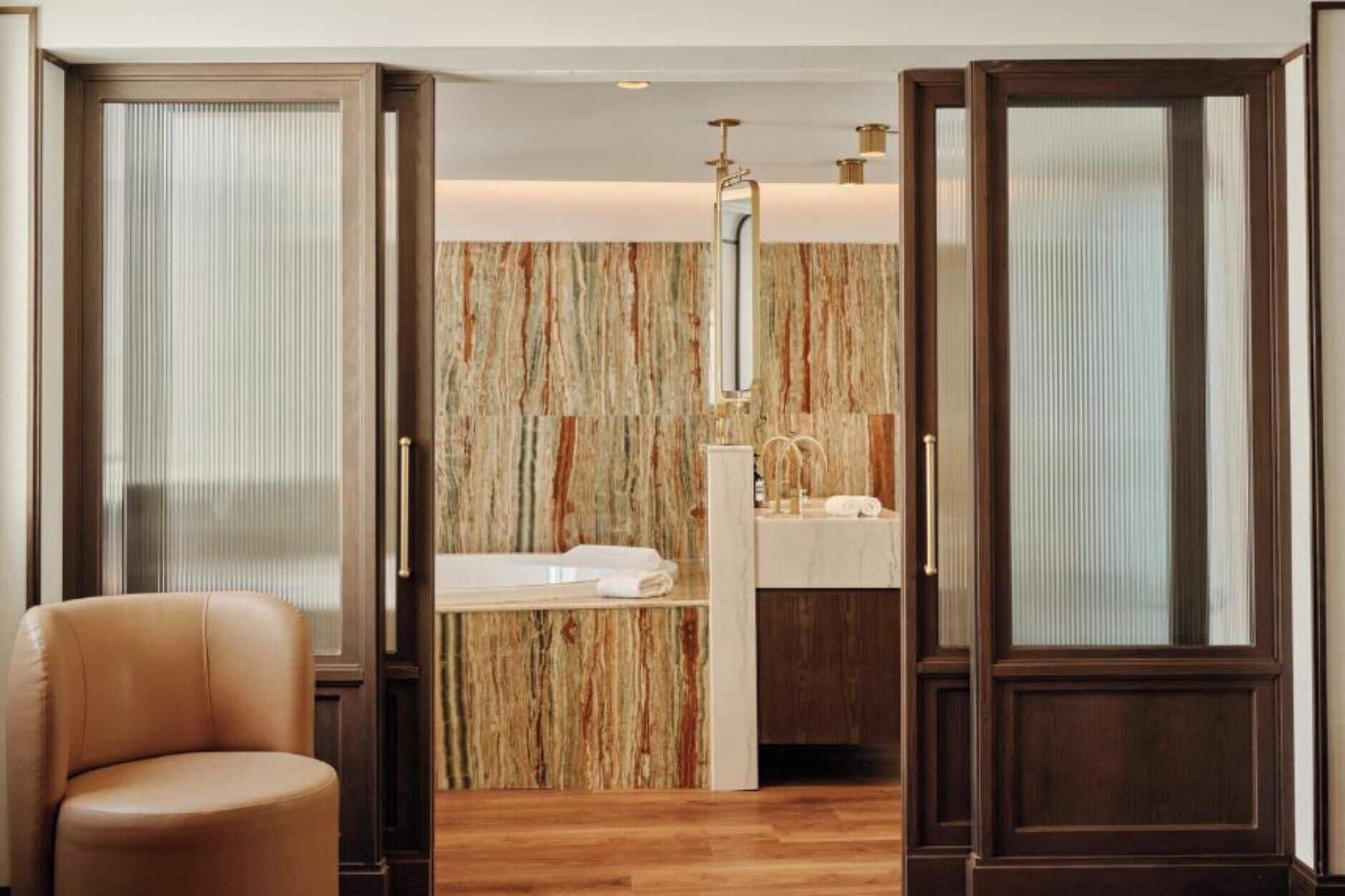 Habitación de hotel con piso de madera, un sillón de cuero color beige, un espejo grande y un baño con pared de mármol y lavamanos.