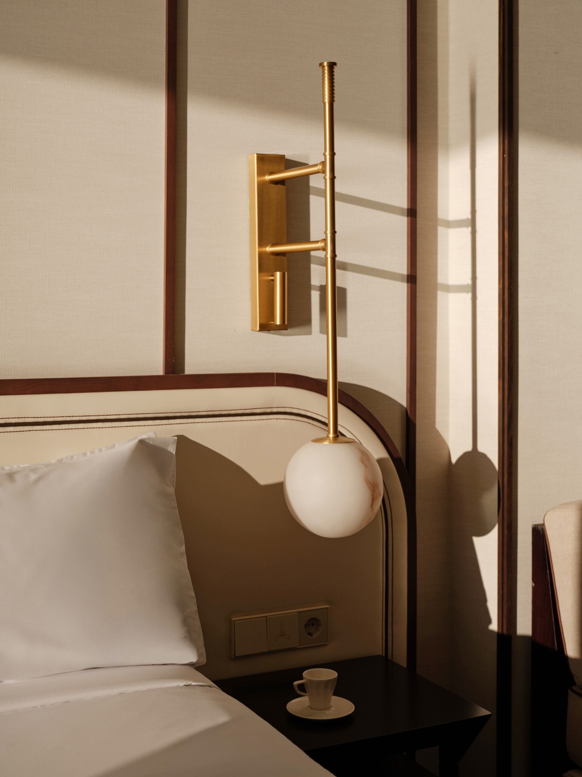 Quarto de hotel com cama, mesa de cabeceira com uma xícara, luminária na parede e tomada.