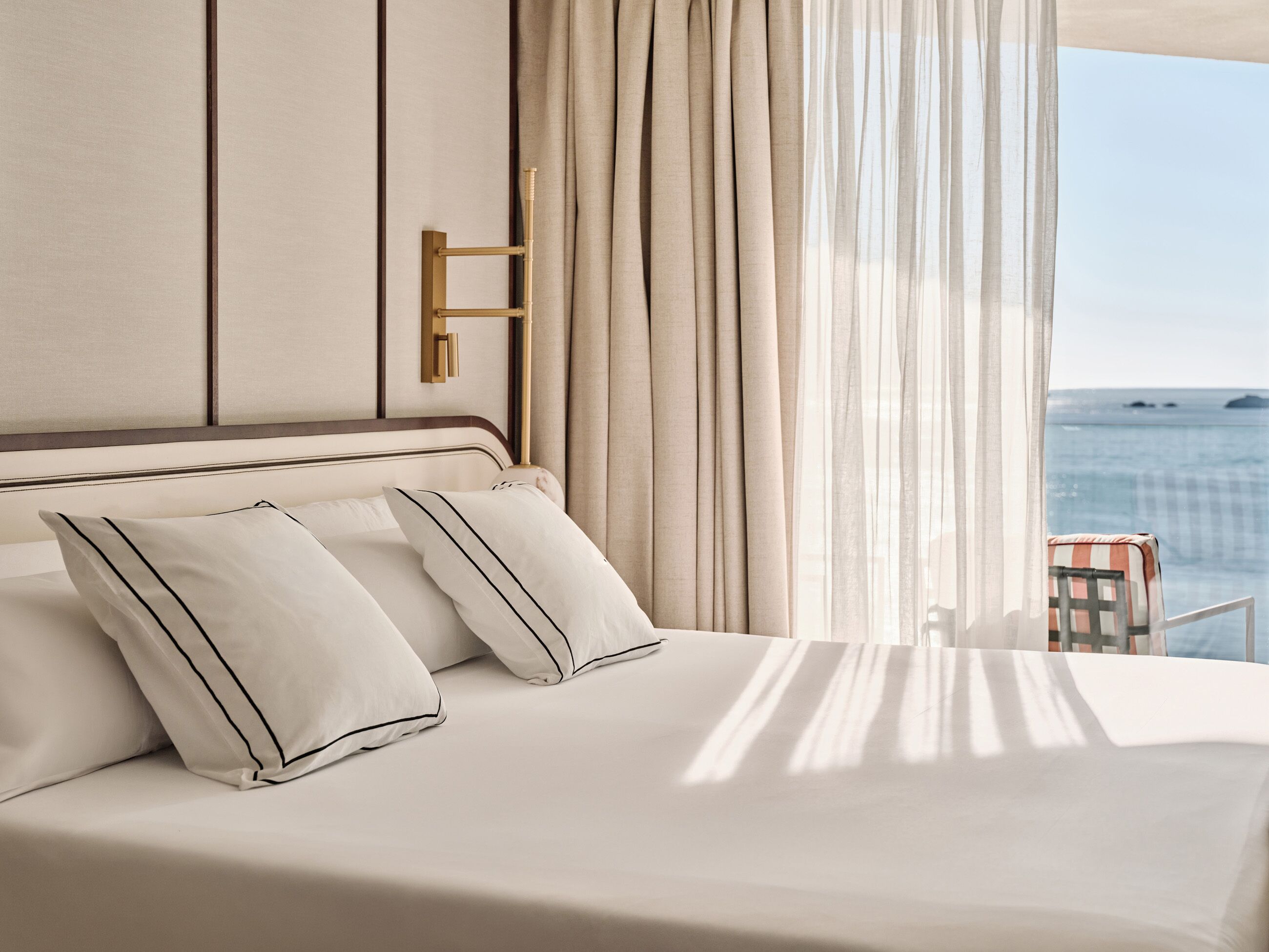 Un lit avec du linge de lit blanc et des bordures noires, une table de chevet dorée, et une vue sur la mer à travers des rideaux transparents.