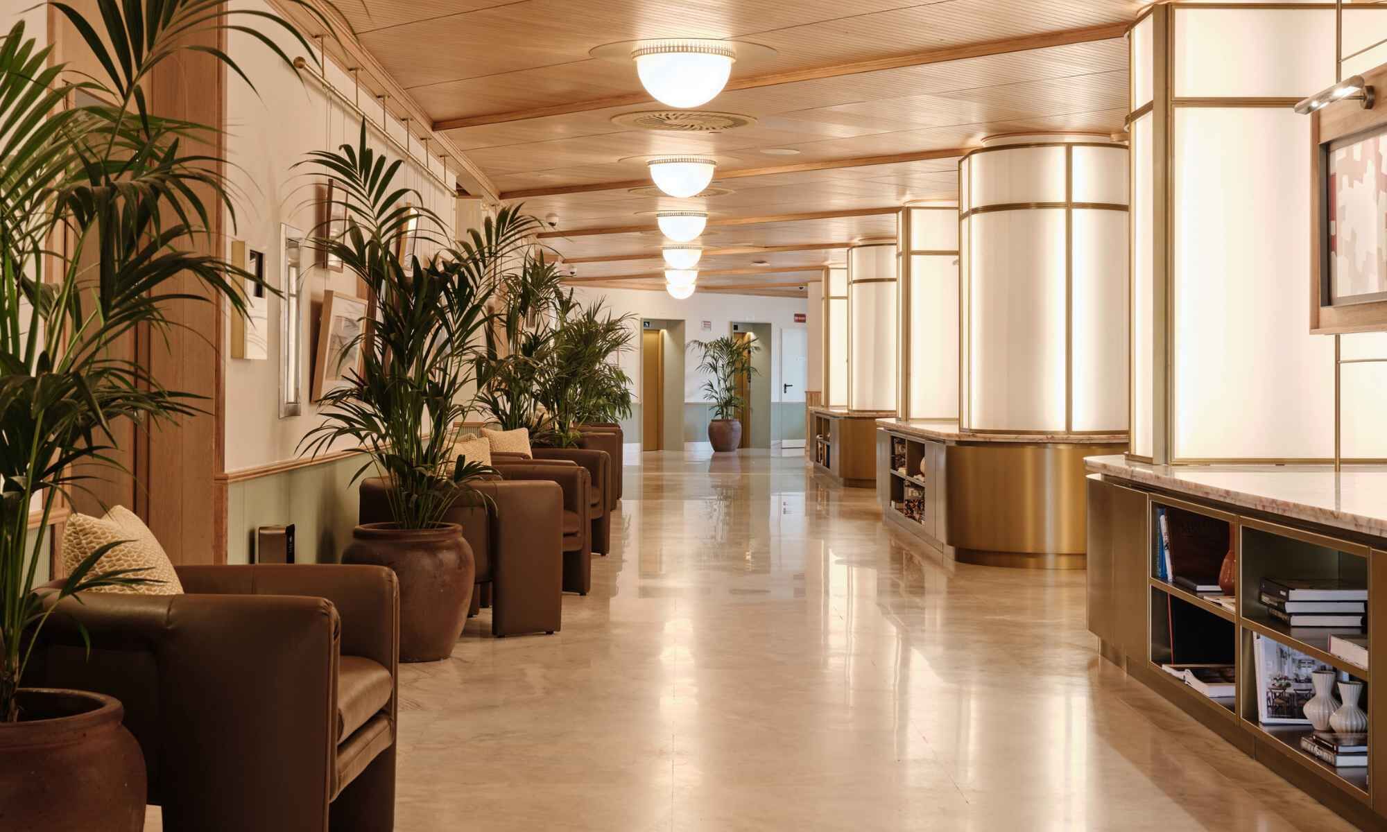 A hotel lobby with a long corridor, leather seating, potted plants, and a reception desk with a bookcase.
