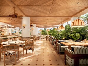 A spacious restaurant with wooden ceiling beams, striped pendant lights, and a tiled floor. Tables are set with white tablecloths and green cushioned chairs. - BLESS Ibiza The Site - New Opening 2026 (Sant Josep de sa Talaia)