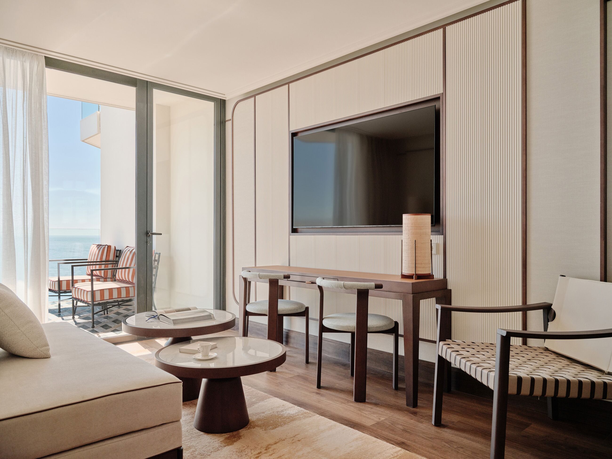 A modern hotel room with a balcony, a TV, a sofa, a dining table, and two chairs.