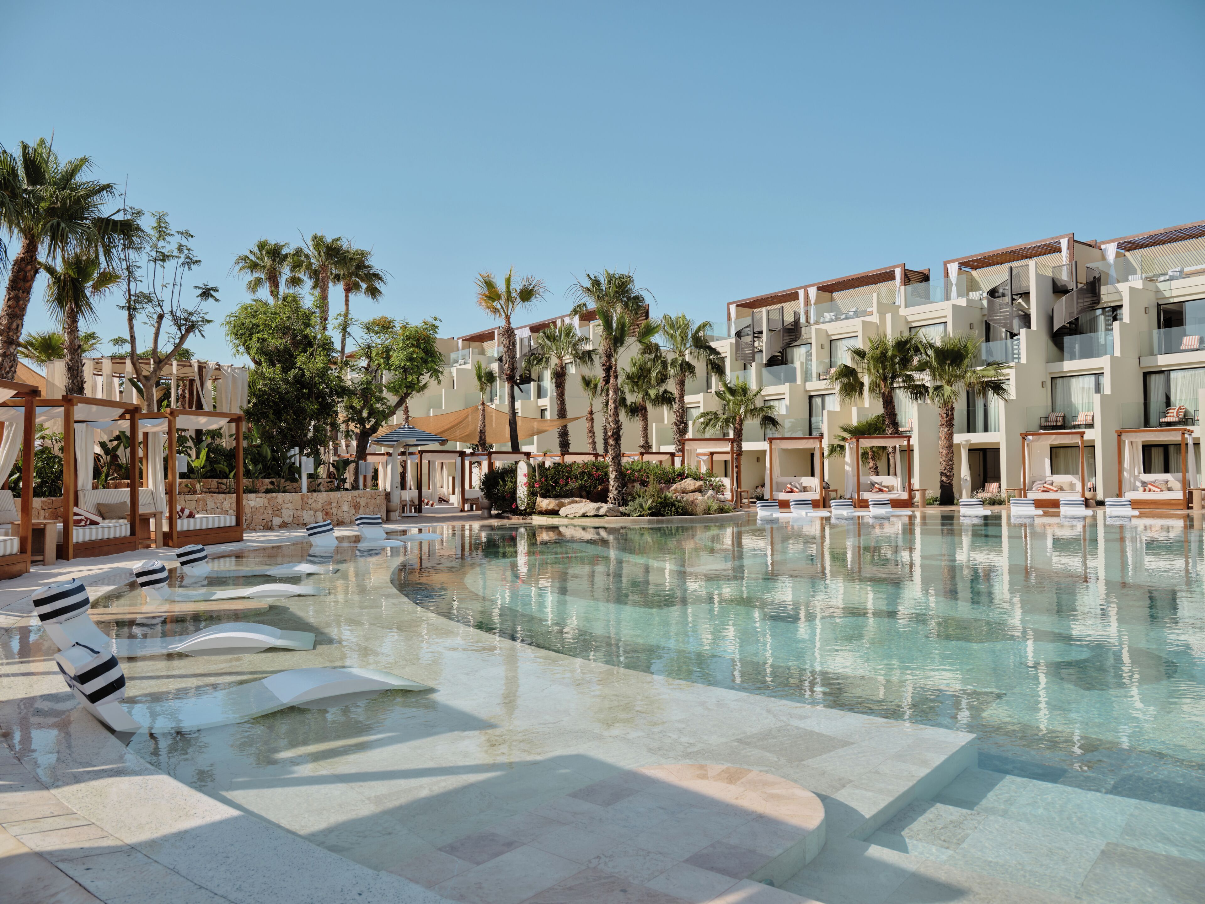 Zona de piscina en un resort con tumbonas, una piscina cristalina y palmeras.
