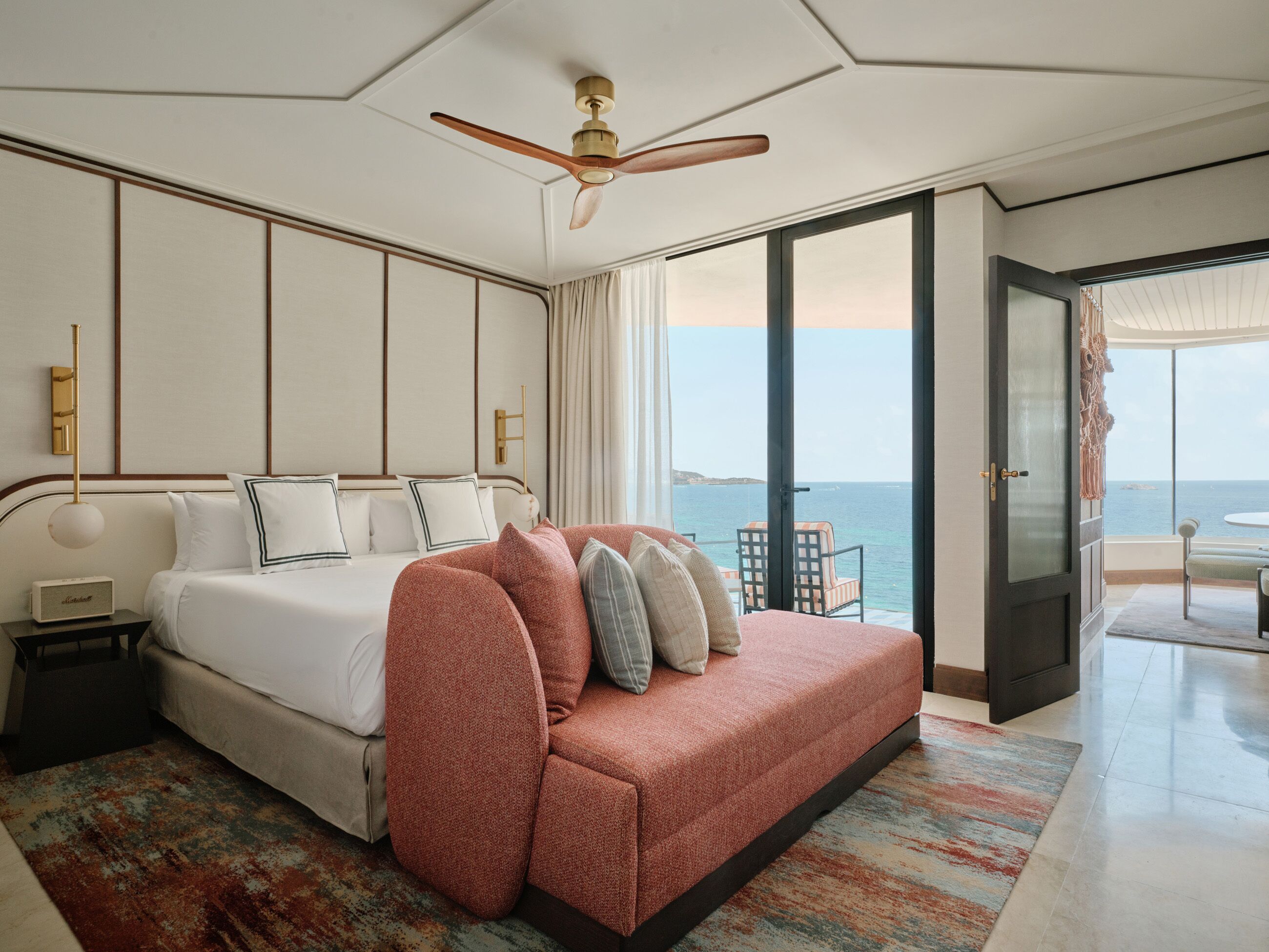A hotel room with a bed, a ceiling fan, a view of the sea, and a balcony with a table and chairs.