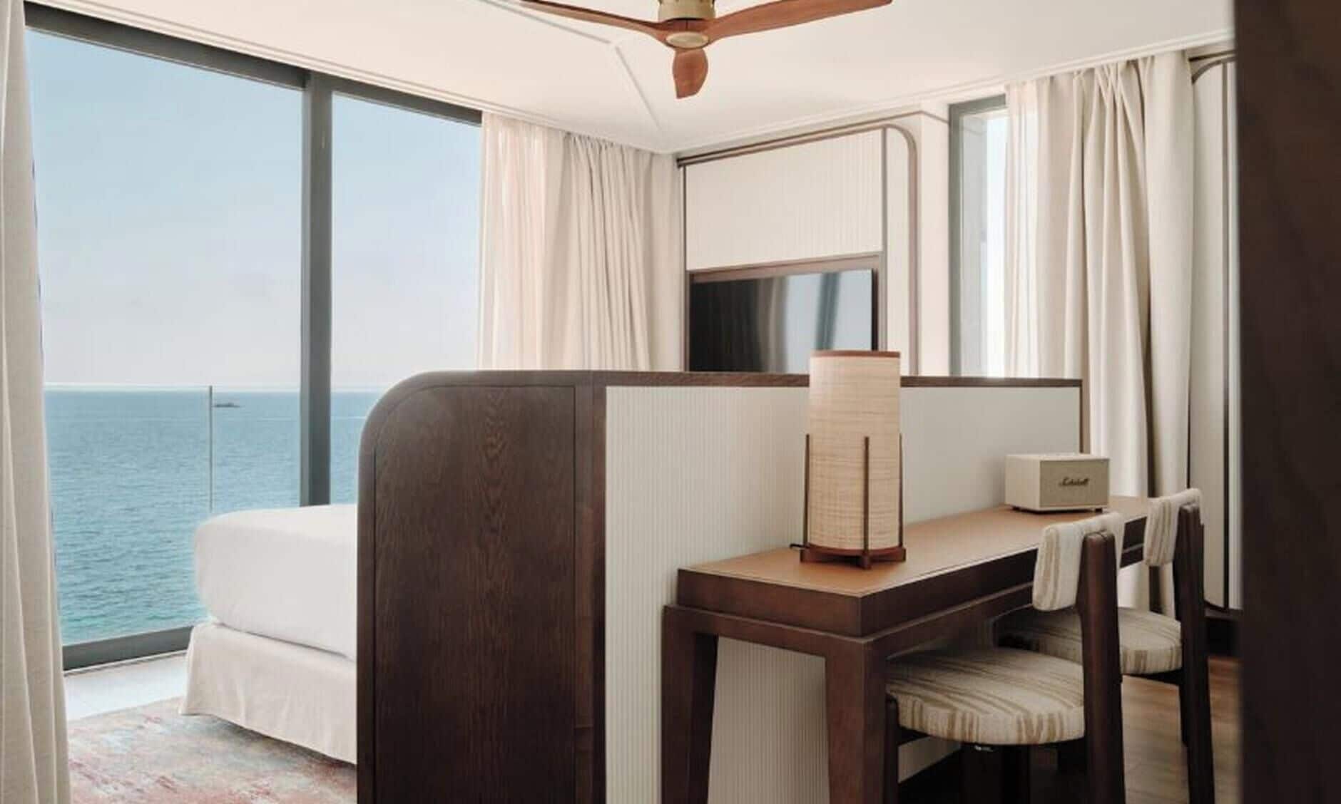 A hotel room with a bed, a desk, a chair, a lamp, and a view of the sea.