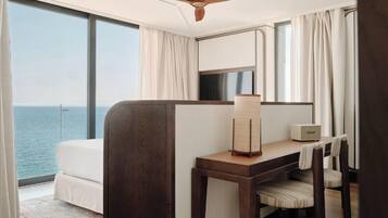A hotel room with a bed, a desk, a chair, a lamp, and a view of the sea.