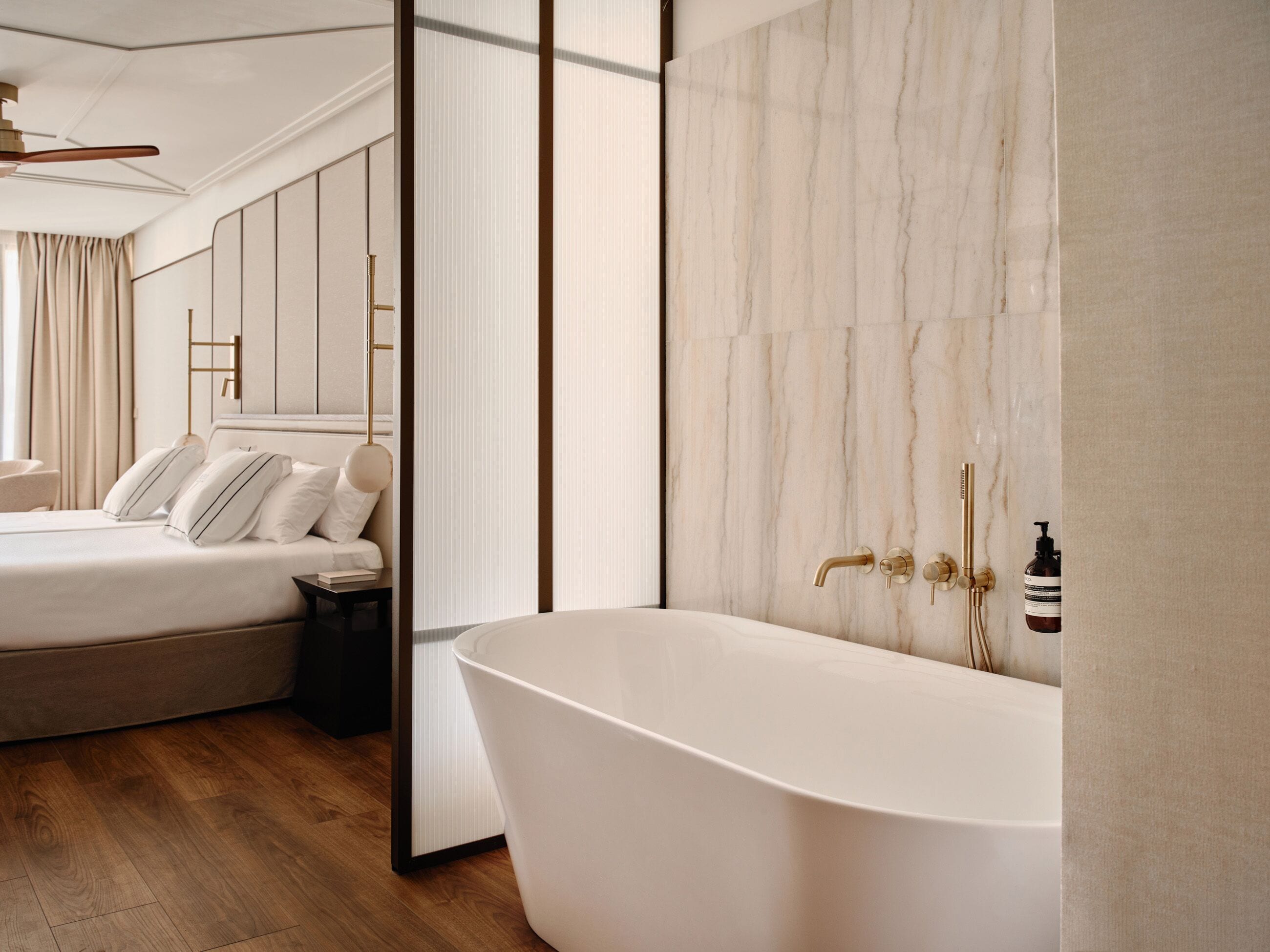 Un dormitorio moderno con una bañera blanca grande, una cama con ropa de cama blanca y piso de madera.