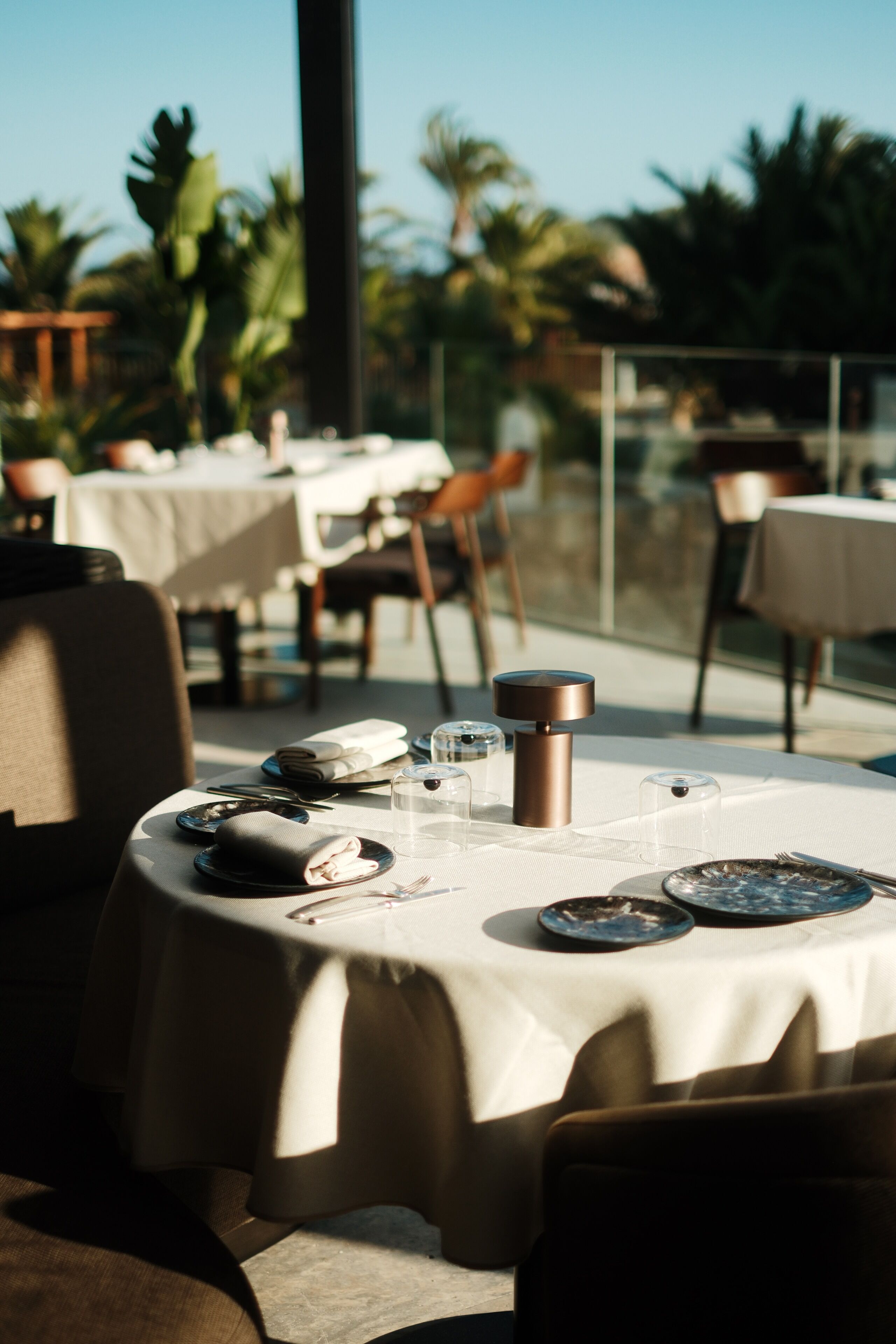 A round dining table with a white tablecloth, set with plates, glasses, and cutlery, surrounded by chairs.
