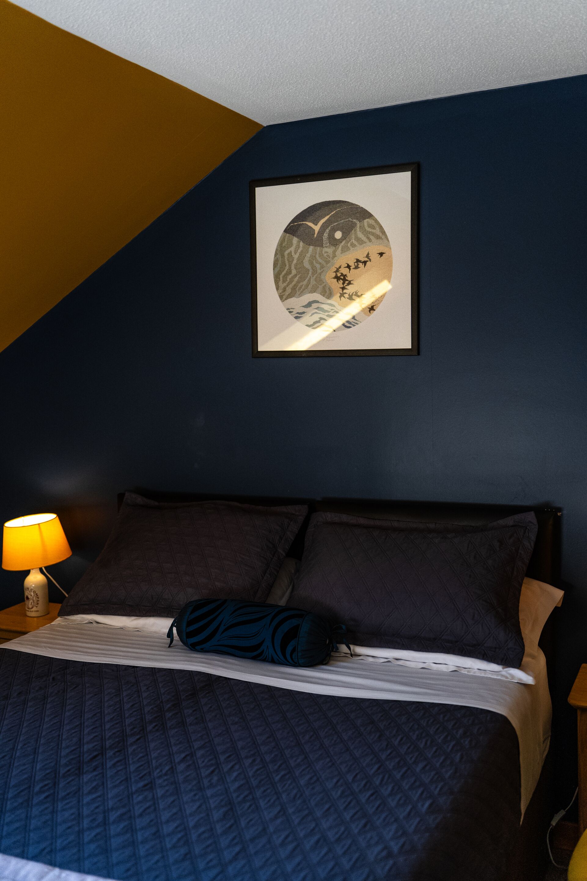 Un dormitorio con cama, almohadas, una obra de arte enmarcada en la pared y una lámpara de noche.