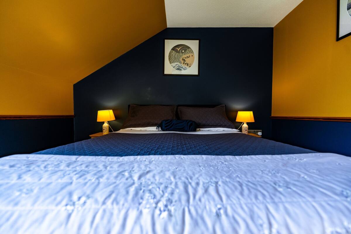 A hotel room with a large bed, two bedside lamps, a framed artwork on the wall, and a dark ceiling.