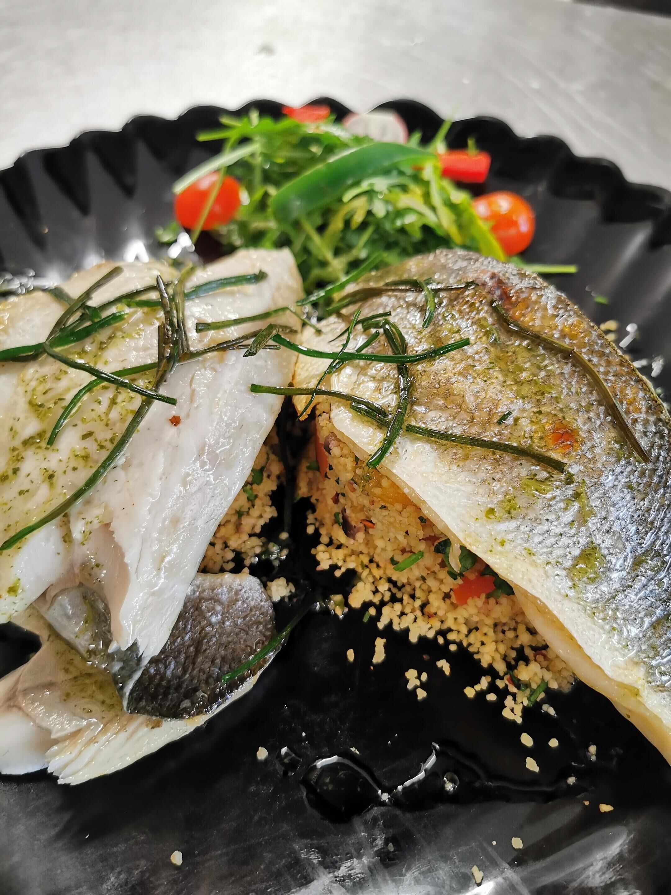 Un plato negro con un filete de pescado, hierbas verdes y cuscús.