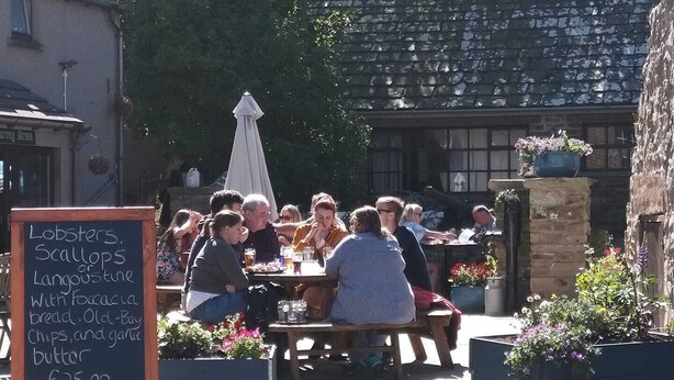An outdoor dining area with people seated at tables, a chalkboard menu, and potted plants.