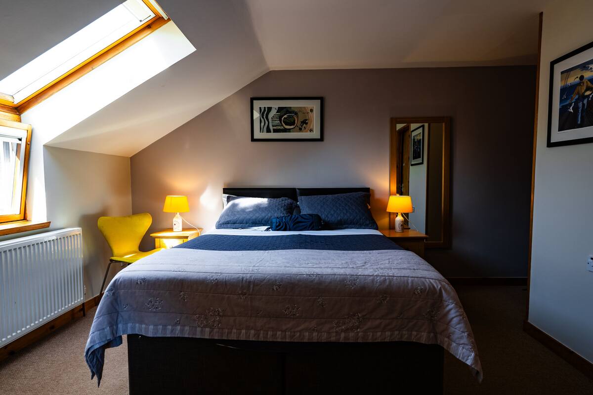 A bedroom with a bed, two bedside lamps, a chair, a window with a view, and a framed picture on the wall.