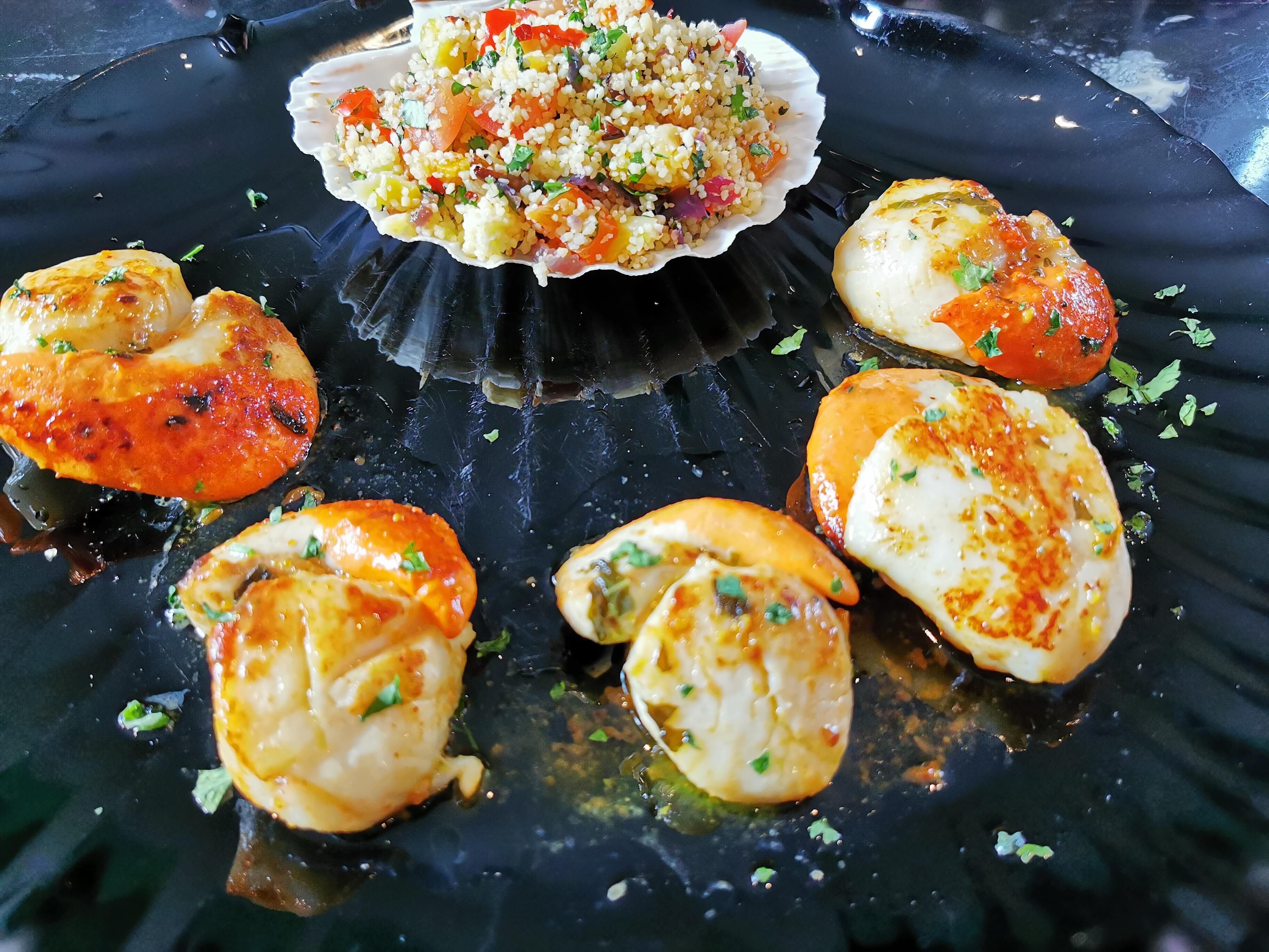 A seafood dish with scallops and a quinoa salad on a black plate.