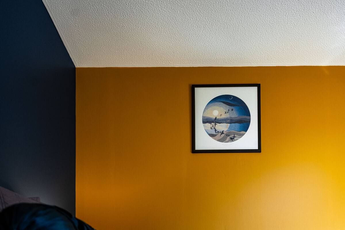 A bed with a blue and white comforter, a dark blue pillow, and a yellow chair against a yellow wall with a framed picture.