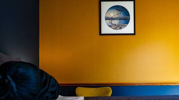 A bed with a blue and white comforter, a dark blue pillow, and a yellow chair against a yellow wall with a framed picture.