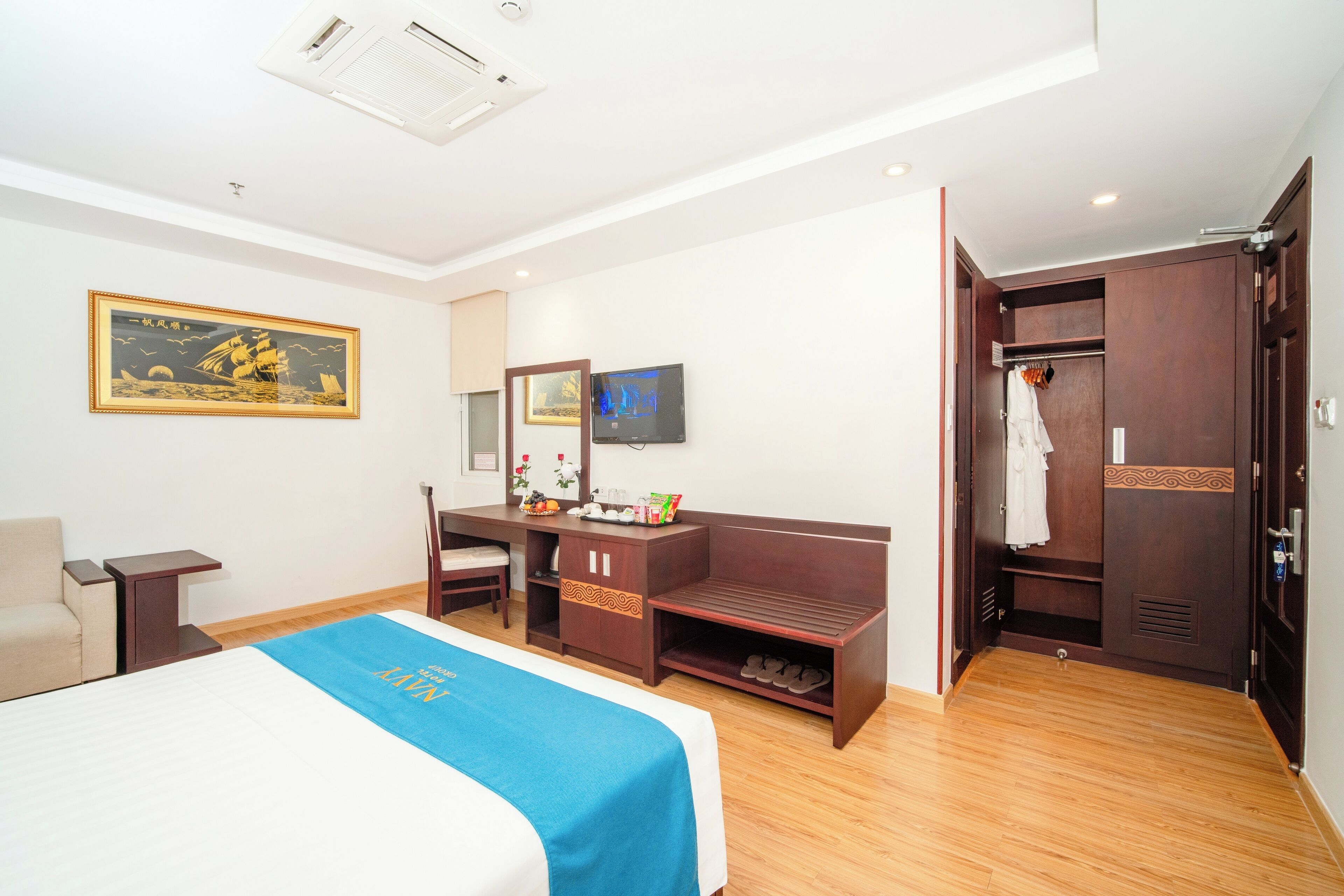 A hotel room with a bed, a desk, a chair, a TV, a wardrobe, and a painting on the wall.