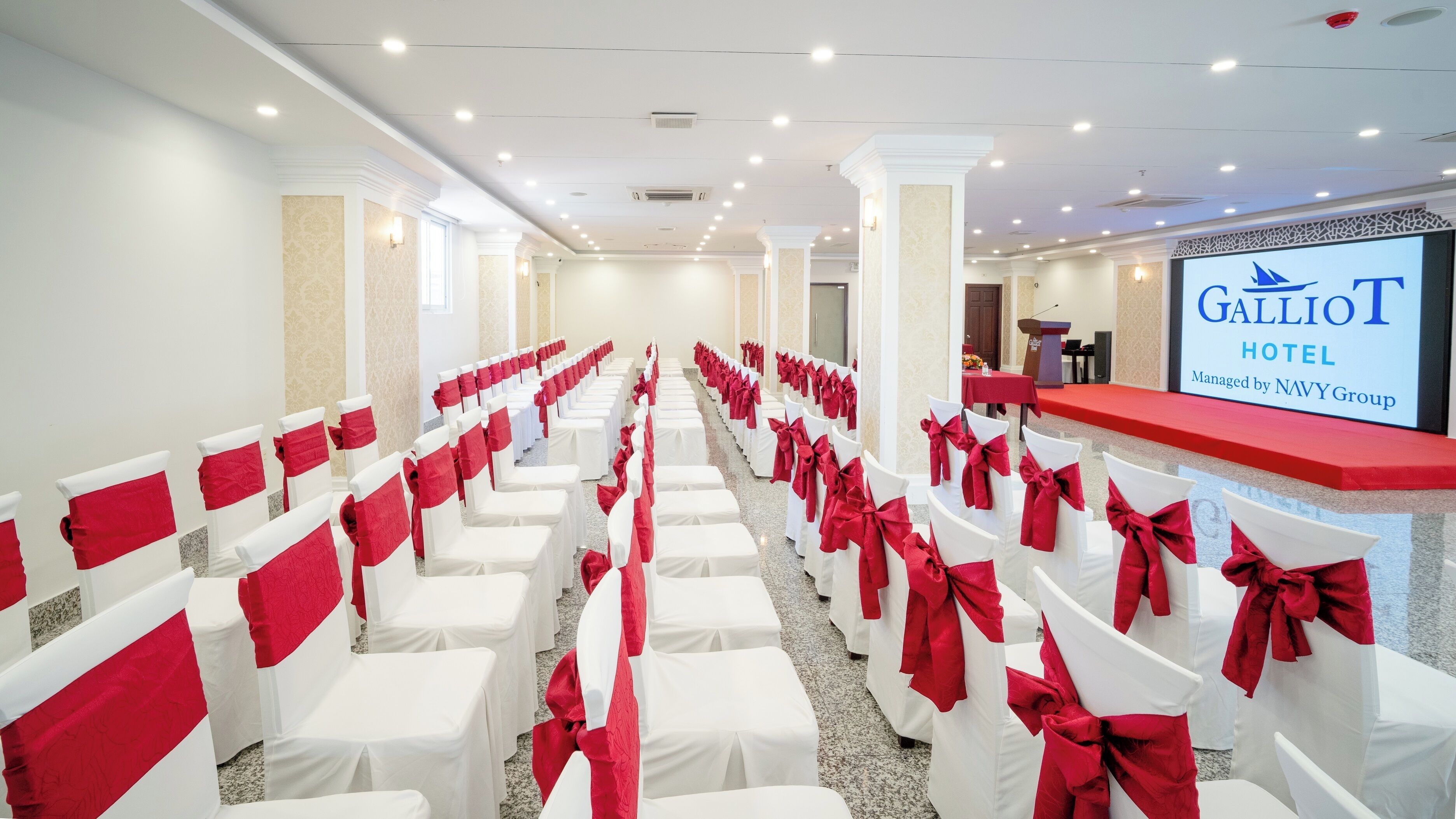 Ein Hotelkonferenzraum mit Reihen weißer Stühle und roten Bändern, einem Rednerpult und einem Bildschirm, der den Namen des Hotels zeigt.