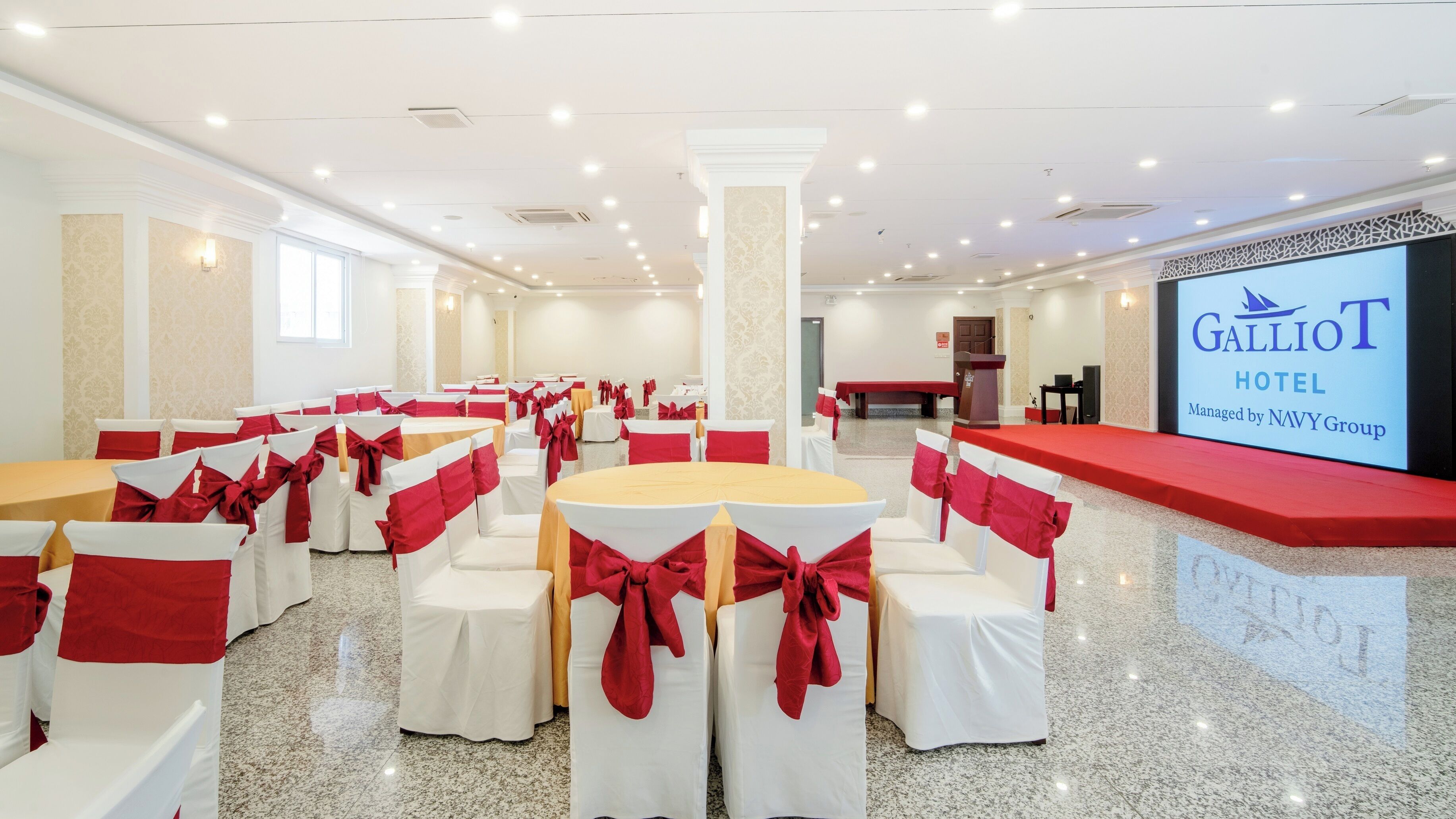 A hotel conference room with round tables and chairs, a stage, and a large screen displaying the hotel name.