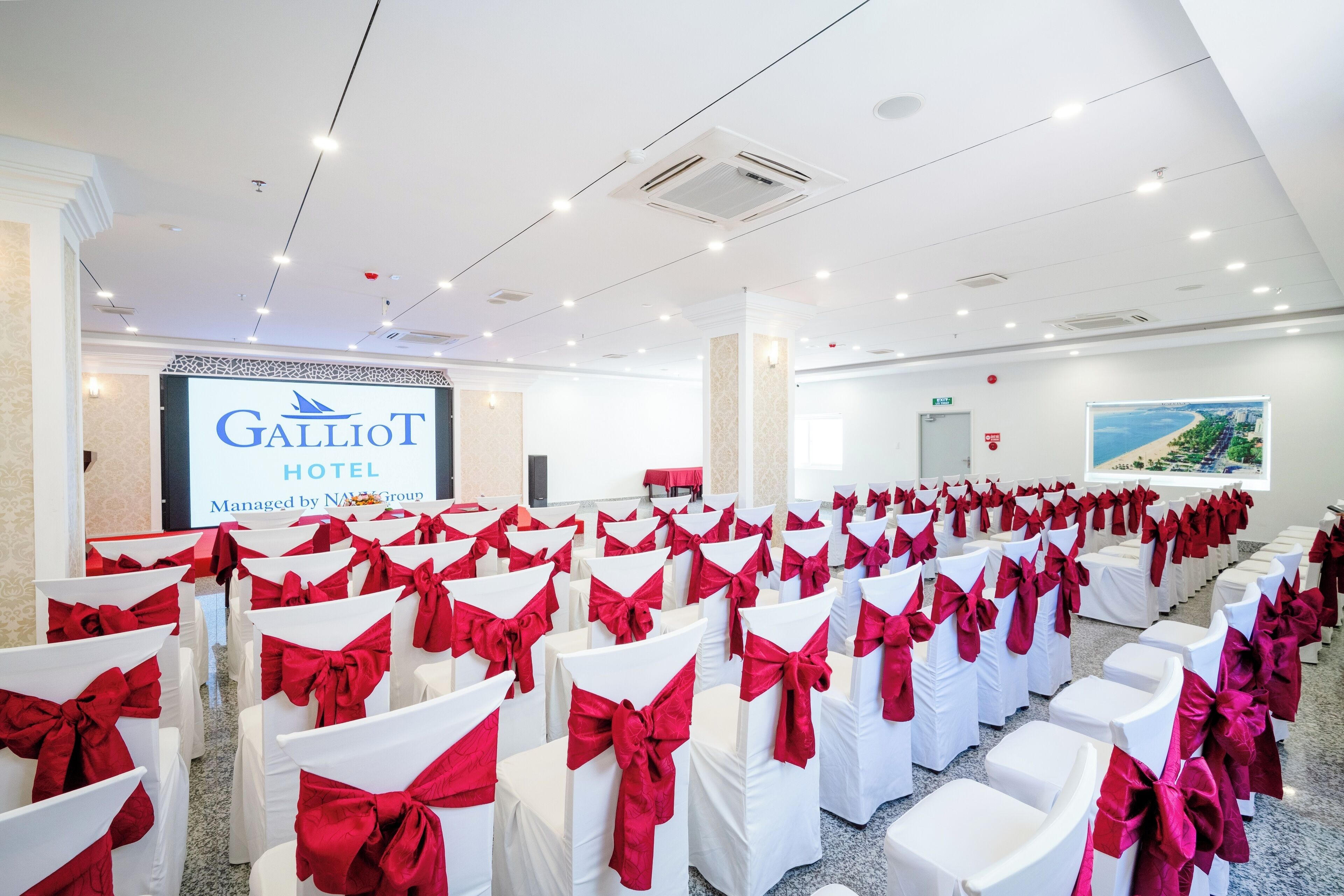 A conference room at Galliot Hotel with rows of white chairs with red sashes, a projection screen displaying the hotel name, and a large window with a view of a coastal landscape.