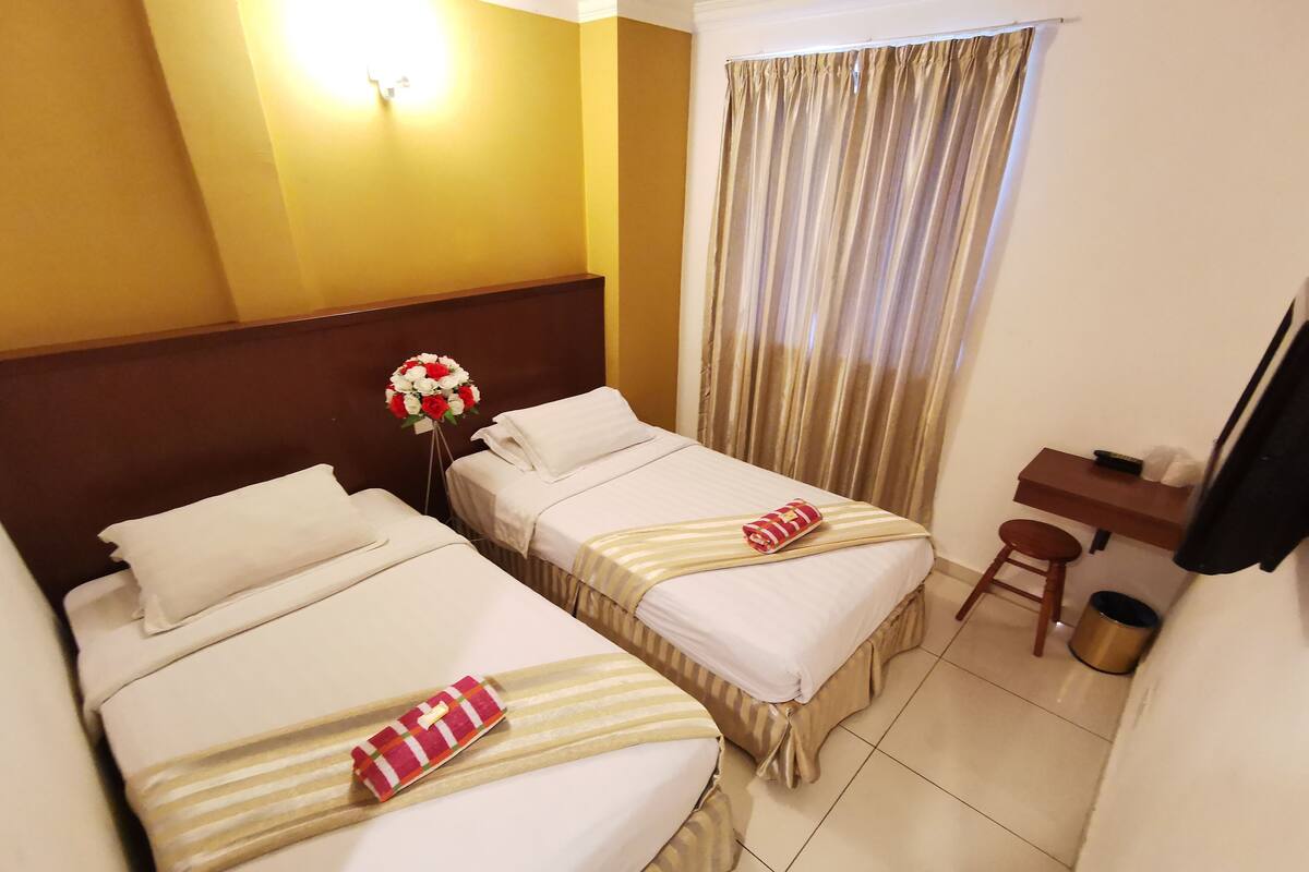 A hotel room with two beds, a desk, a chair, a trash can, and a wall-mounted TV.