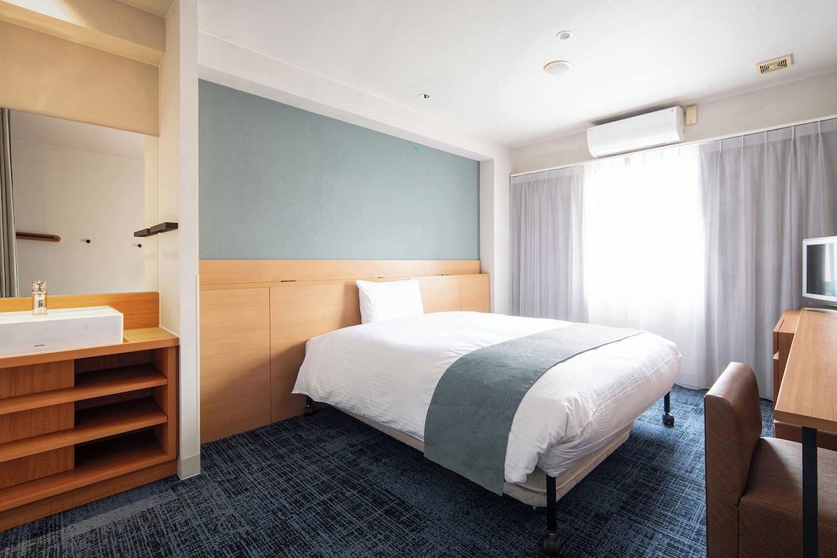 A hotel room with a bed, a desk with a TV, a bathroom with a sink and mirror, and a window with curtains.
