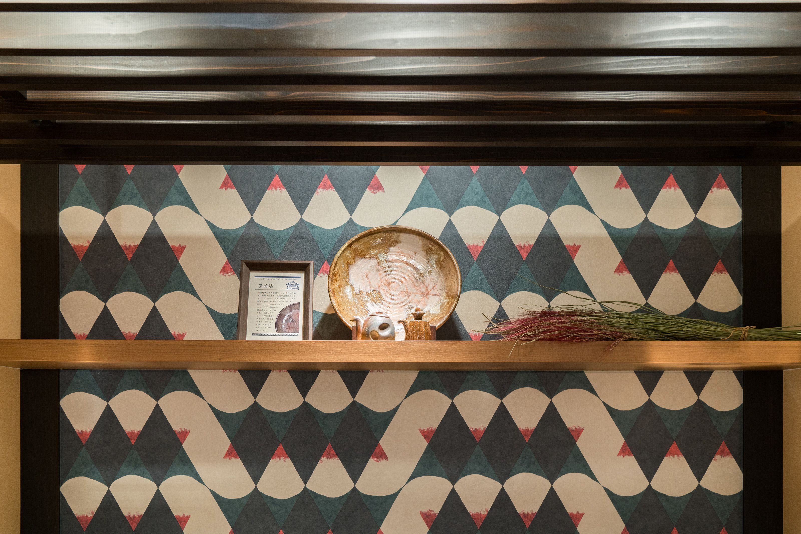 A patterned wall with a shelf displaying a teapot, a framed certificate, and a dried plant.