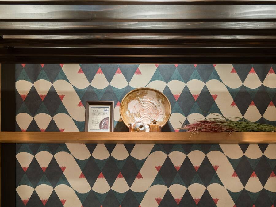 Un mur orné d’un motif décoratif, sur lequel est posée une étagère où l’on peut voir une théière, un certificat encadré et une plante séchée.