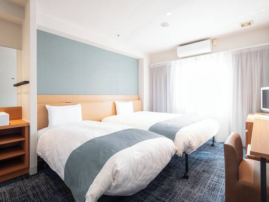 A hotel room with two beds, a desk, a TV, a bathroom with a sink, and a large window with curtains.