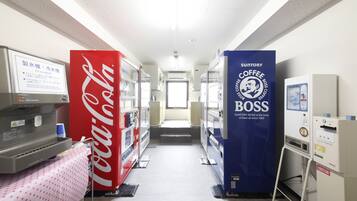 Un couloir où se trouvent des distributeurs automatiques, notamment un distributeur de Coca-Cola et un autre de café de la marque Suntory Coffee Boss, ainsi qu’une table recouverte d’une nappe à pois.