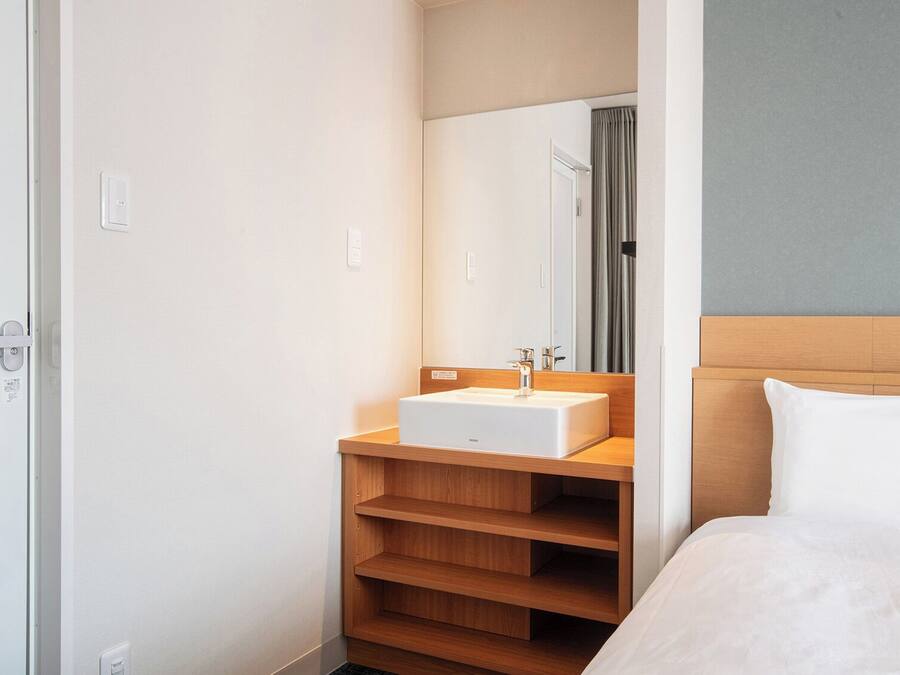 A hotel room with a bed, a wooden shelf unit, a white sink, and a mirror.