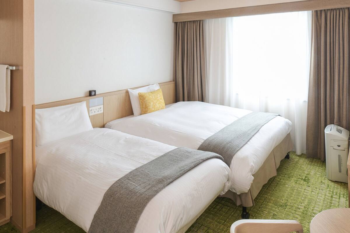 Habitación de hotel con dos camas, una mesita de noche, una silla y una ventana con cortinas.